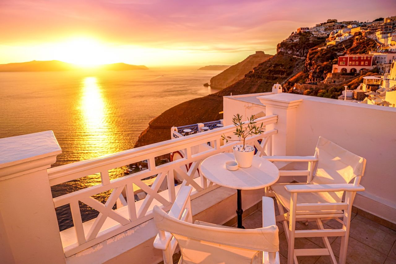 Idyllic Santorini sunset from a patio overlooking the caldera, with white chairs and table.